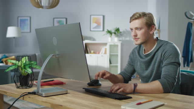 Portrait-of-the-Handsome-Man-Working-on-Personal-Computer-while-Sitting-at-His-Desk.-In-the-Background-Stylish-Cozy-Living-Room.-Young-Freelancer-Working-From-Home.