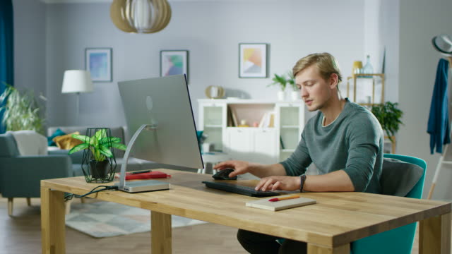 Retrato-de-la-centra-joven-hombre-trabajando-en-un-ordenador-mientras-está-sentado-en-su-escritorio.-En-el-fondo-acogedora-sala-de-estar.