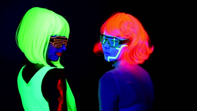 Women-with-UV-face-paint,-wig,-glowing-glasses,-glowing-clothing-dancing-together-slowly-in-front-of-camera,-Half-body-shot.-Caucasian-and-asian-woman.-.