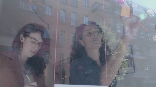 Two-businesswomen-brainstorm-on-windows-wall-over-real-estate