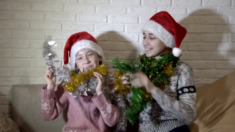 Cheerful,-happy-little-girls-in-red-hats-of-Santa-Claus-play-with-each-other-tinsel,-laugh-and-smile-sitting-on-the-couch-against-a-white-brick-wall.