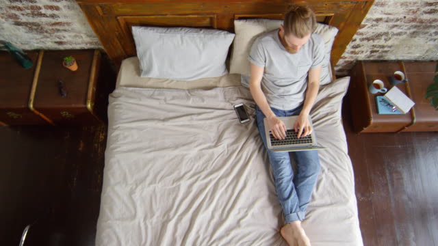 Junger-Mann-sitzen-auf-Bett-und-Arbeit-am-Laptop