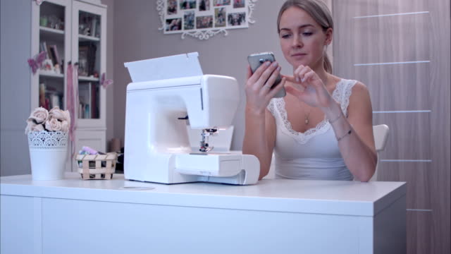 Young-woman-with-phone-sitting-at-the-table-next-to-sewing-machine