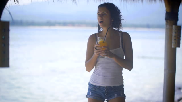 Sexy-brunette-in-white-tank-top-and-jeans-shorts-is-dancing-near-the-sea-in-tropical-bar-enjoying-her-vacation.-Beautiful-young-girl-is-relaxing-with-cocktail-on-the-beach-moving-with-the-music