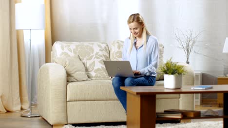 Smiling-Young-Woman-Working-on-a-Laptop.-She's-in-Her-Living-Room-Sitting-on-a-Sofa-and-Holding-Notebook-on-Her-Lap.