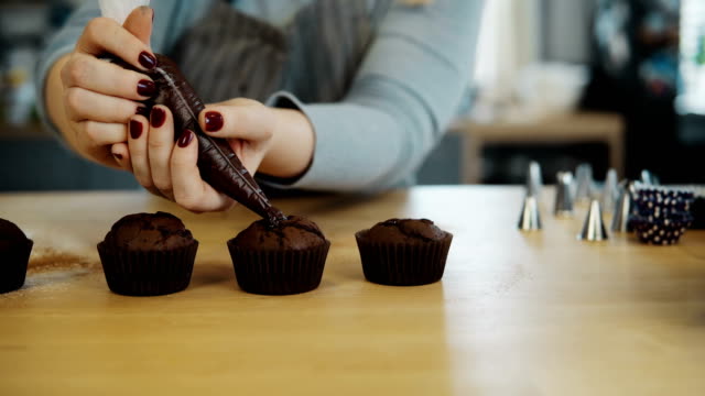 Close-up-view-of-female-hands-puts-the-cream-from-pastry-bag-on-chocolate-cupcakes.-Young-woman-cooking-the-muffins