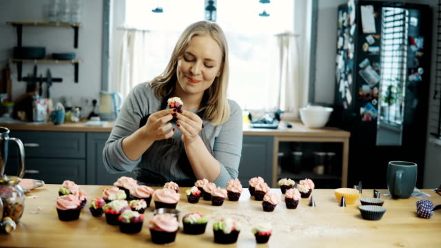 Young-woman-standing-on-the-kitchen-and-tasting-chocolate-cupcakes-with-cream-on-the-top.-Confectioner-prepares-muffins