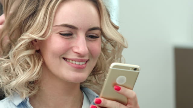Mujer-con-peinado-en-el-salón-mientras-navega-por-internet-en-su-teléfono-móvil,-sonriendo
