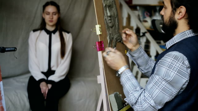 Sculptor-creating-sculpture-of-human's-face-on-canvas-while-young-woman-posing-him-in-art-studio