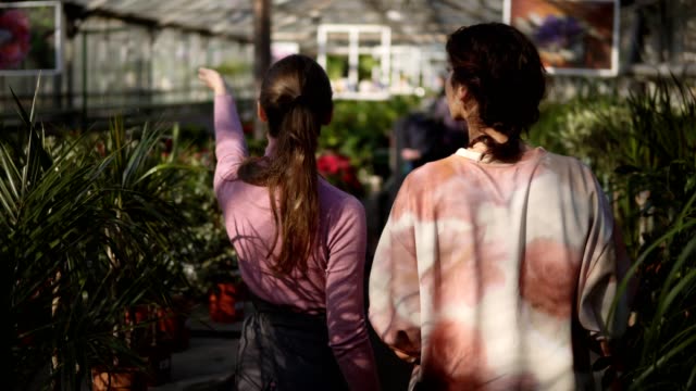Back-view-of-young-female-florist-walking-with-a-client-and-showing-her-different-plants,-explaining-information.-Young-woman-is-listening-carefully-to-the-florist