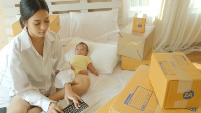 Joven-madre-asiática-y-su-niño-recién-nacido,-comprobación-orden-del-ordenador-portátil-para-el-cliente-y-entrega-en-línea-de-embalaje-listo-en-dormitorio.
