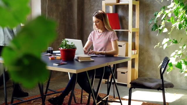 Young-woman-is-working-with-laptop-sitting-at-table-in-office.-Her-colleague-is-coming,-women-start-watching-screen-together-and-laughing.-Informal-friendly-atmosphere.