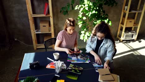 High-angle-view-of-young-designers-are-placing-photos-on-table-to-create-perfect-flat-lay-working-at-studio.-They-are-sitting-at-table-together-and-discussing-different-photos