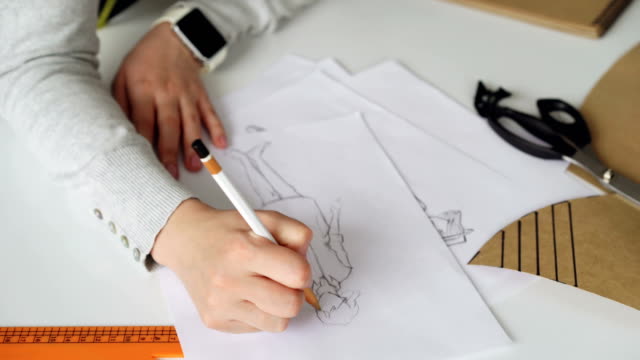 Tilt-up-of-Fashion-designer-is-drawing-lines-on-clothing-sketch-with-coffee,-tailor's-scissors,-ruler-and-pieces-of-paper-on-studio-table.-Attractive-girl-is-busy-and-concentrated-on-work.