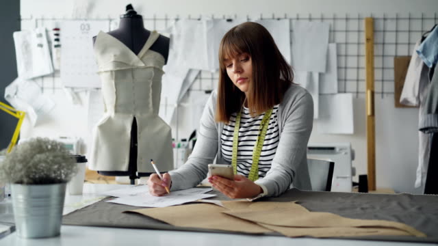 Kreative-Modedesigner-ein-Blick-auf-Smartphone-und-Skizze-während-der-Arbeit-in-modernen-Schneiderei-am-nähtisch.-Frau-ist-beschäftigt-und-Prozess-beteiligt.