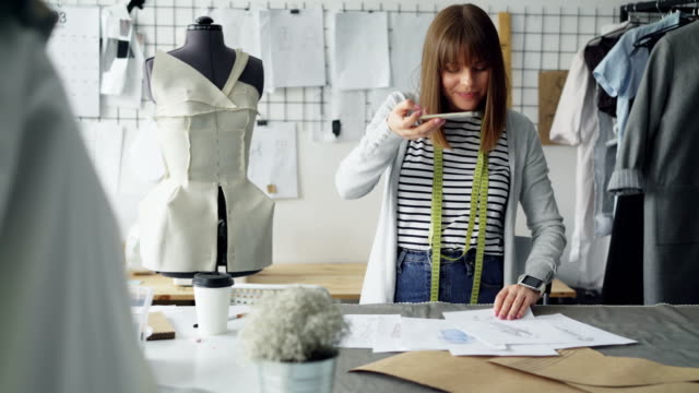 Ambitious-creative-female-tailor-is-placing-garment-sketches-on-studio-desk-and-shooting-them-with-smartphone.-Presenting-drawings-of-new-collection-concept.