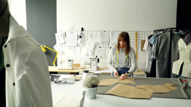 Young-attractive-female-designer-is-checking-her-paper-templates-with-measuring-tape.-Professional-creative-clothing-designing-concept.-Studio-is-light-and-modern.