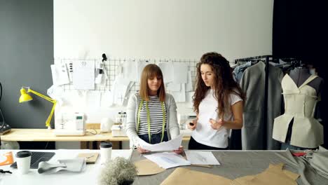 Emprendedores-de-diseño-de-moda-joven-están-discutiendo-los-bocetos-de-la-nueva-colección-de-ropa-en-su-estudio-de-la-luz.-Las-mujeres-buscan-a-través-de-dibujos,-gesticulando-y-hablando.