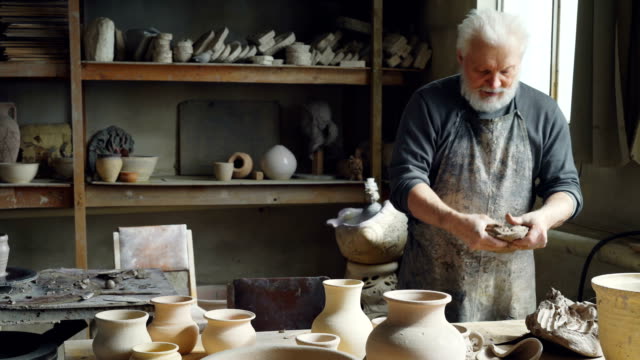 Professional-male-ceramist-is-kneading-clay,-forming-clay-ball-while-working-in-small-workshop-with-potter's-equipment,-tools-and-many-ceramic-figures.