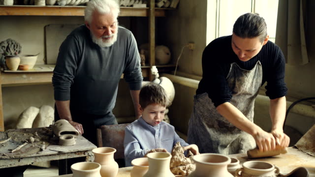 Professional-potter-is-kneading-clay-on-worktable-in-home-studio-while-his-son-is-helping-him-and-his-elderly-father-watching-them-from-behind.-Small-family-business-concept.