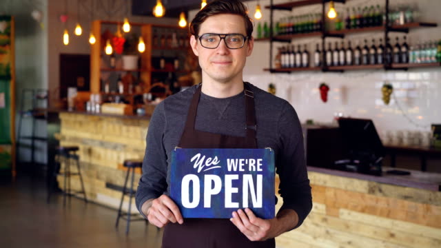 Portrait-of-cheerful-handsome-young-waiter-in-apron-holding-"we-are-open"-sign-standing-in-opening-coffee-shop.-Starting-business-and-successful-people-concept.