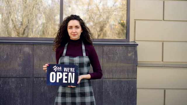 Portrait-of-attractive-confident-woman-in-apron-small-business-owner-holding-"yes-we-are-open"-sign-standing-outside-and-smiling-looking-at-camera.