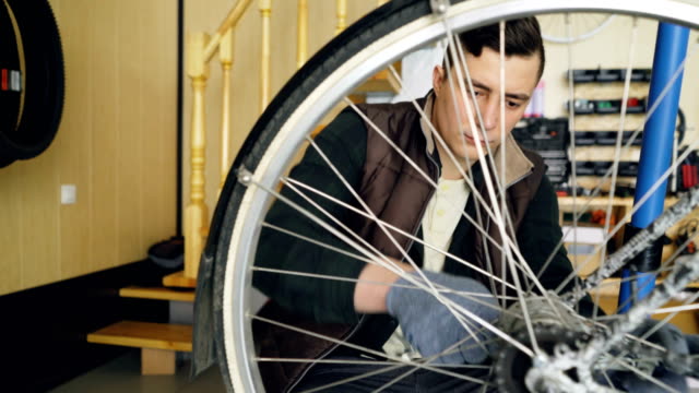 Young-man-experienced-serviceman-is-fixing-bike-wheel-using-wrench-and-tools.-Small-cozy-workshop-interior-with-wooden-walls-and-ladder,-spare-parts-and-equipment-are-visible.