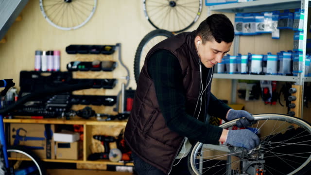 Handsome-mechanic-is-cleaning-bicycle-wheel-outside-and-inside,-hub-and-spokes-with-piece-of-cloth-and-listening-to-music-with-earphones.-Profession-and-people-concept.