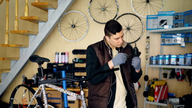 Konzentrierte-Mechaniker-ist-Mechanismus-während-der-Reparatur-Fahrrad-in-schönen-Workshop-einfetten.-Junger-Mann-in-warme-Weste-und-Schutzhandschuhe-hört-Musik.