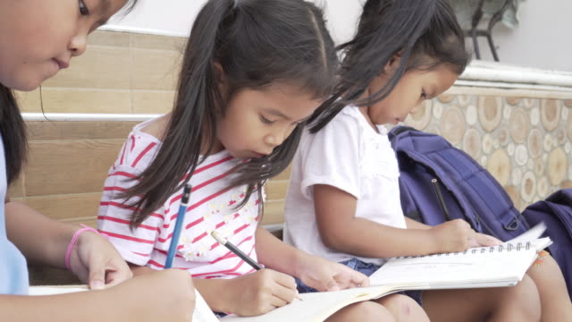 Alumno-asiático-dibujar-en-los-cuadernos-de-rodillas-durante-una-clase-al-aire-libre