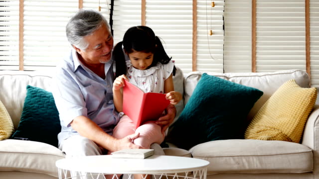 Senior-man-and-little-girl-reading-book-together-at-home.-people-with-family,-lifestyle,-education-concept.-4k-resolution.