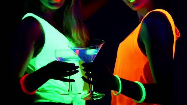 Women-with-UV-face-paint,-laser,-glowing-bracelets,-drinks,-glowing-clothing-dancing-together-in-front-of-camera,-Mid-shot.-Caucasian-and-asian-woman.-.