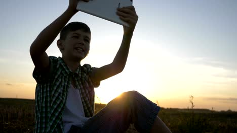 niño-se-sienta-en-la-parte-superior-y-habla-en-la-comunicación-de-vídeo-utiliza-una-tableta,-al-aire-libre