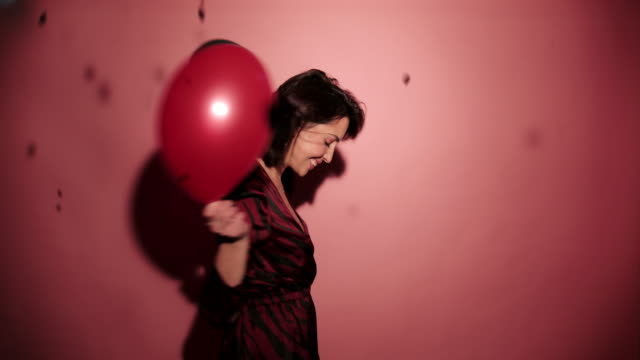 Brunette-happy-woman-dance-with-balloons-and-confetti-in-pink-background-wear-red-dress