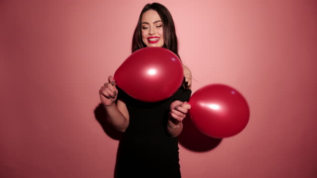 Brunette-happy-woman-dance-with-balloons-and-confetti-in-pink-background-wear-red-dress