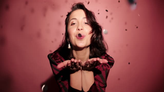 Brunette-happy-woman-give-a-kiss-to-camera-with-confetti-in-pink-background-wear-red-dress