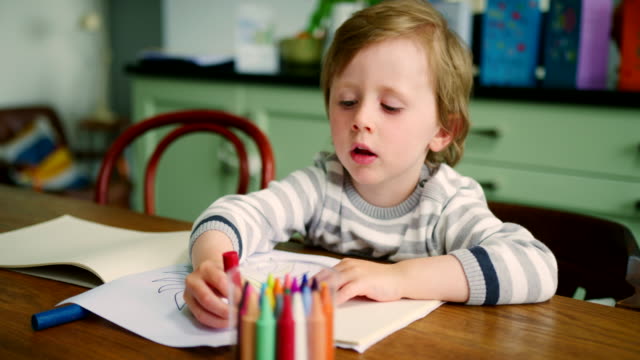 Joven-niño-rubio-sentado-en-la-mesa-de-la-cocina-usando-lápices-de-colores