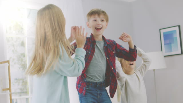 Two-Cute-Little-Girls-and-Young-Adorable-Boy-Have-Fun,-Jumping-High-on-a-Couch-at-Home.-Happy-Kids-Dancing-on-a-Sofa-in-the-Sunny-Living-Room.-In-Slow-Motion.