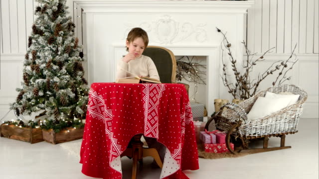 Chico-sonriente-explorando-las-puturas-en-el-libro-sentado-en-la-mesa-cerca-del-árbol-de-Navidad