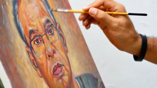 Closeup-artist-hand-holding-paint-brush-painting-portrait-of-asian-man