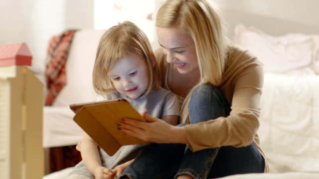 Schöne-Mutter-und-Ihre-kleine-Tochter-haben-gute-Zeit-beim-Lesen-von-Kinderbüchern-auf-einem-Tablet-Computer.