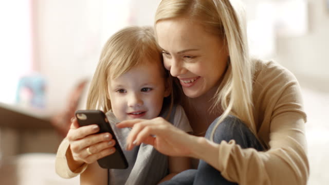 Schöne-junge-Mutter-setzt-sich-mit-Ihrer-kleinen-Tochter-durch-und-zeigt-Ihr-Interessantes-auf-einem-Smartphone.-Das-Kinderzimmer-ist-rosa-und-voll-mit-Spielzeug.