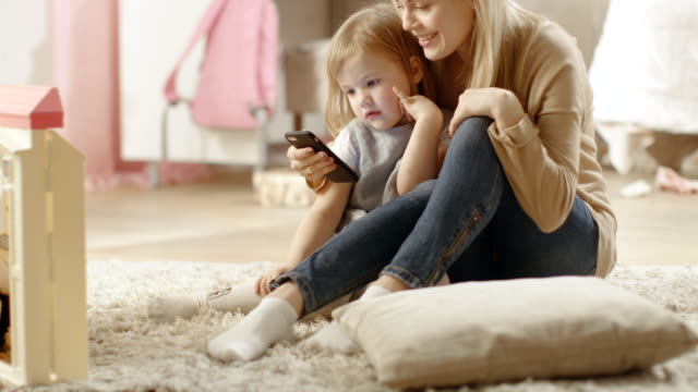Beautiful-Young-Mother-Sits-with-Her-Little-Daughter-and-Shows-Her-Something-Interesting-on-a-Smartphone.-Children's-Room-is-Pink-and-Full-of-Toys.
