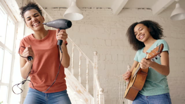 Mixed-race-young-funny-girls-dance-singing-with-hairdryer-and-playing-acoustic-guitar-on-a-bed.-Sisters-having-fun-leisure-in-bedroom-at-home