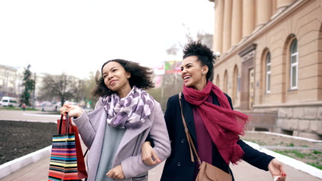 Dolly-shot-of-two-attractive-mixed-race-women-dancing-and-have-fun-while-walking-down-the-street-with-shopping-bags.-Happy-young-friends-walk-after-visiting-mall-sale
