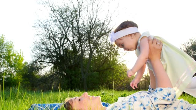 love-is-above-all,-In-the-park,-on-the-grass,-on-the-lawn,-lying-down,-mom,-a-young-blond-woman-playing,-smiling,-with-her-daughter,-a-little-cute-baby,-in-the-sun,-at-sunset.-Im-having-fun,-mom-raising-up-baby,-baby-laughing