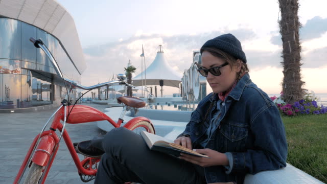 Joven-mujer-leyendo-un-libro-con-bicicleta-vintage-al-lado-en-primera-línea-de-mar-al-amanecer