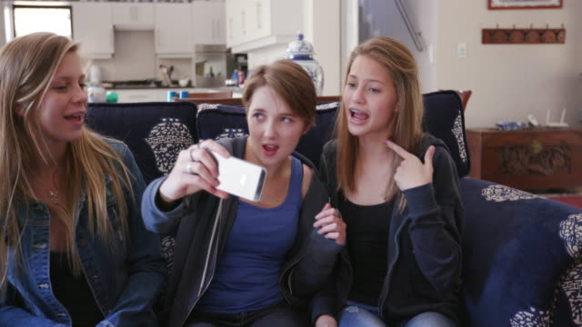 Three-pretty-young-teenage-girls-sitting-on-a-sofa,-taking-a-selfie-video-and-having-fun