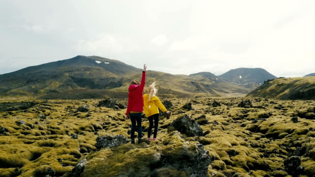 Aerial-view-of-two-woman-standing-on-the-rock-in-lava-field-in-Iceland.-Tourists-looks-on-landscape,-dancing-and-jumping