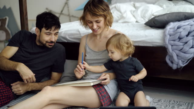 Familia-feliz-con-la-hija-adorable-Linda-dibujo-en-el-álbum-con-lápices-sentado-junto-a-la-cama-en-casa